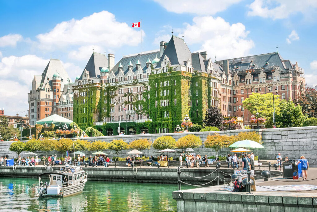 A large, ivy-covered historic hotel with a Canadian flag rises over a lively waterfront in one of the best boating cities Canada offers, bustling with people, boats, and market stalls.