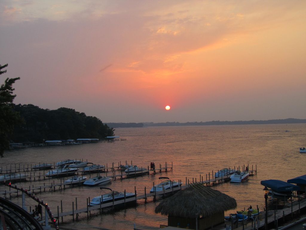 A calm view of West Lake Okoboji at sunset glowing under an orange sky. Boats are docked at wooden piers, and a thatched-roof hut sits in the foreground, inviting you to relax after a long day on the water.