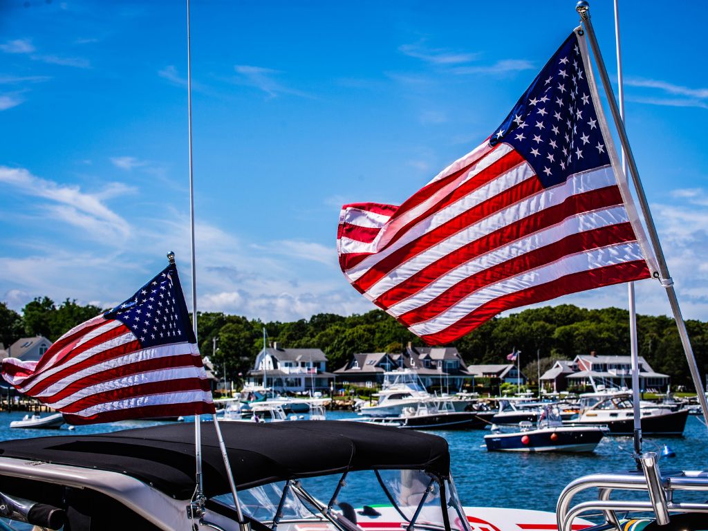Two American flags wave from boats in the foreground at one of the best Labor Day boating destinations. Waterfront houses line the shore, and more boats glide across the water under a clear blue sky.