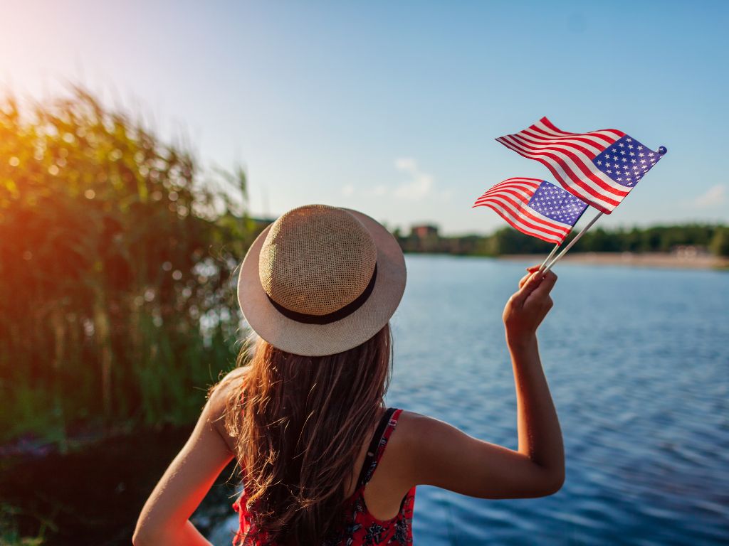 A person wearing a hat stands by a lake, holding two small American flags, with sunlight and greenery all around.