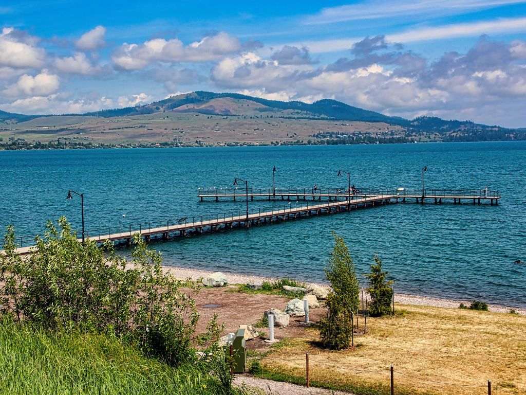 A wooden pier extends into a blue lake with mountains and clouds behind. The shoreline in front is a mix of grass and sand, perfect for relaxing before or after your day on the water.