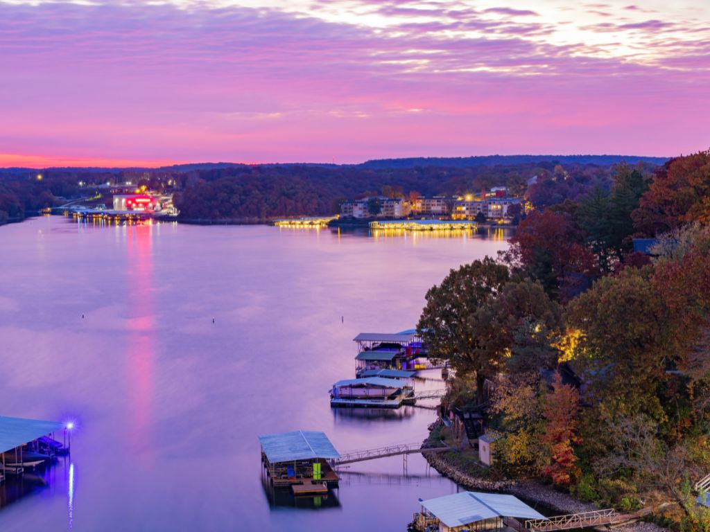 An aerial view of the calm waters of Lake of the Ozarks at sunset. You’ll see docks and boats scattered along the shore, with colorful buildings and trees reflected beneath a stunning sky painted in purple, pink, and orange hues.
