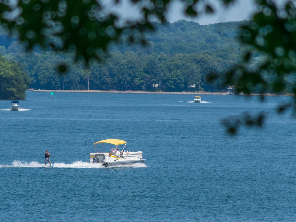A view from among the trees towards a person wakeboarding behind a pontoon boat with a yellow canopy on a Smith Mountain Lake, one of the best Labor Day boating destinations. Other boats and leafy tree branches frame the scene, highlighting the lively atmosphere.