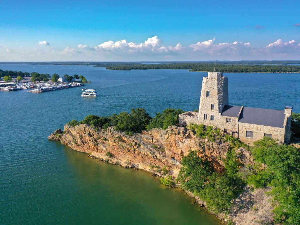 A stone building with a tall tower sits on a rocky peninsula overlooking one of the best Labor Day boating destinations in Lake Murray, SC. There's a boat passing by, and you can see a marina under a blue sky.