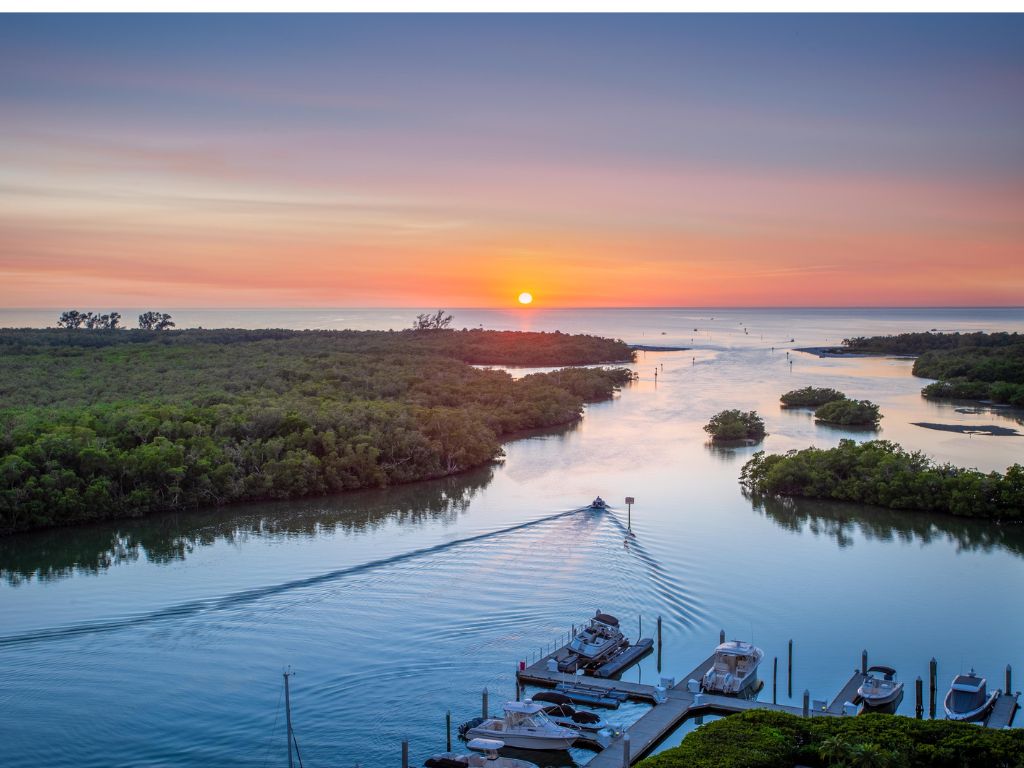 A small boat glides through a calm waterway at sunset, surrounded by green islands, it's one of the best Labor Day boating destinations. Boats are docked nearby, and the sky beyond glows orange.