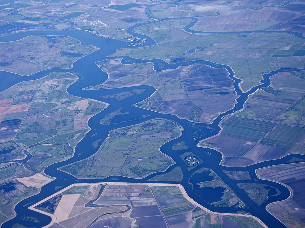 An aerial view of a river delta shows winding waterways, patches of farmland, and glimpses of late summer boating along the water channels. 