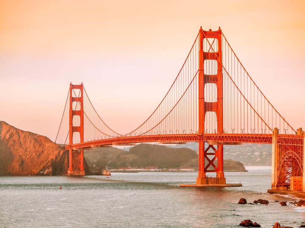 The Golden Gate Bridge stretches across the water with hills behind it, captured under a warm, orange-tinted sky during late summer boating. 