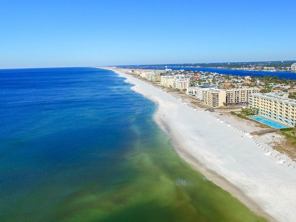 An aerial view of a sandy beach lined with mid-rise buildings and a calm blue ocean, perfect for late summer boating under a clear sky.