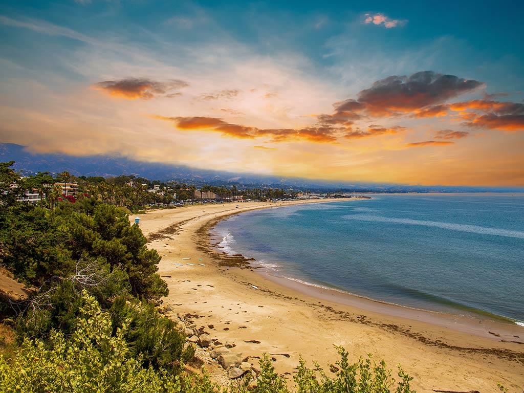 A wide sandy beach curves along the shoreline, bordered by greenery and distant buildings, as late summer boating unfolds beneath a dramatic sunset sky with orange clouds.