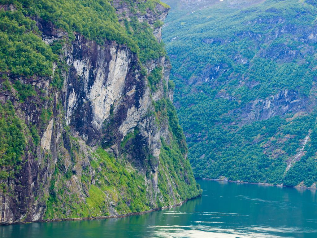 Steep, rocky cliffs covered in green vegetation rise above a calm, narrow body of water in a fjord landscape in Norway.