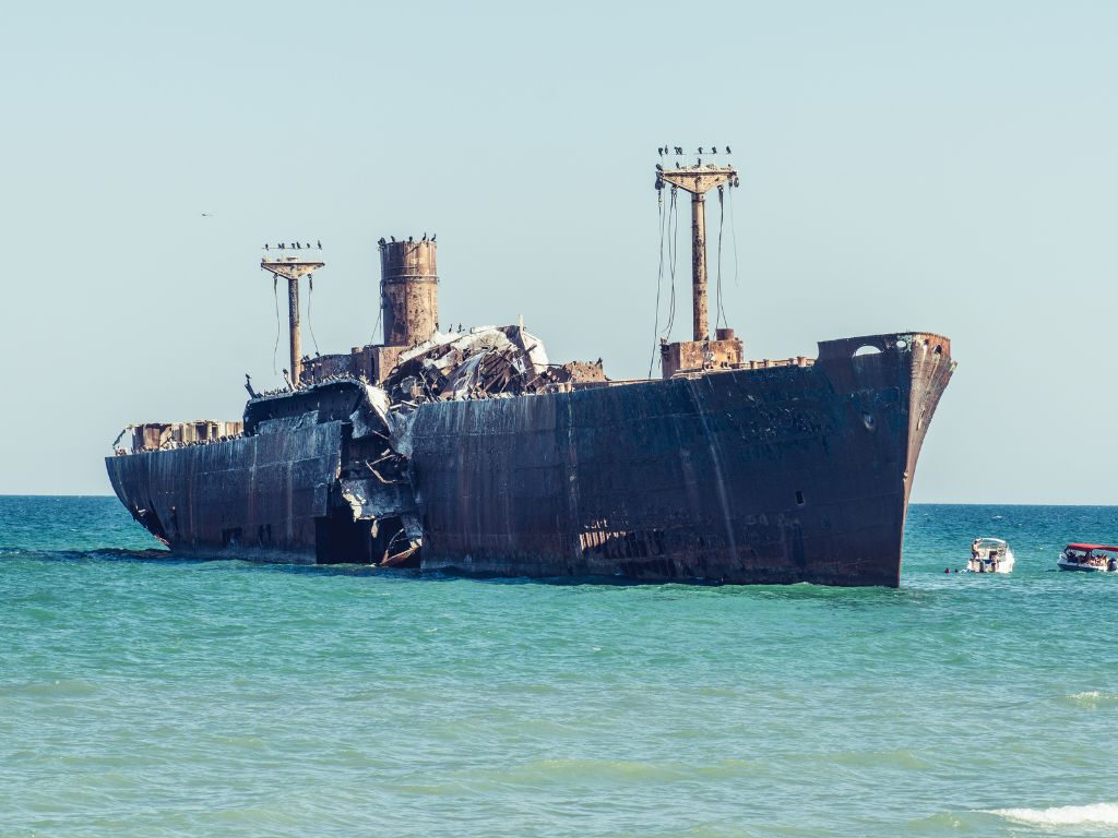 A large, rusted shipwreck's partially submerged in shallow ocean water, with several birds perched on its masts and small boats nearby — one of the spookiest boating destinations in Europe.