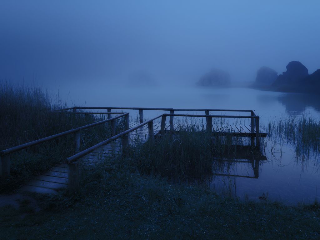 A wooden dock stretches into a calm, misty lake with tall grasses and distant rocks, evoking the eerie charm of Europe’s spookiest boating spots in blue-toned light.
