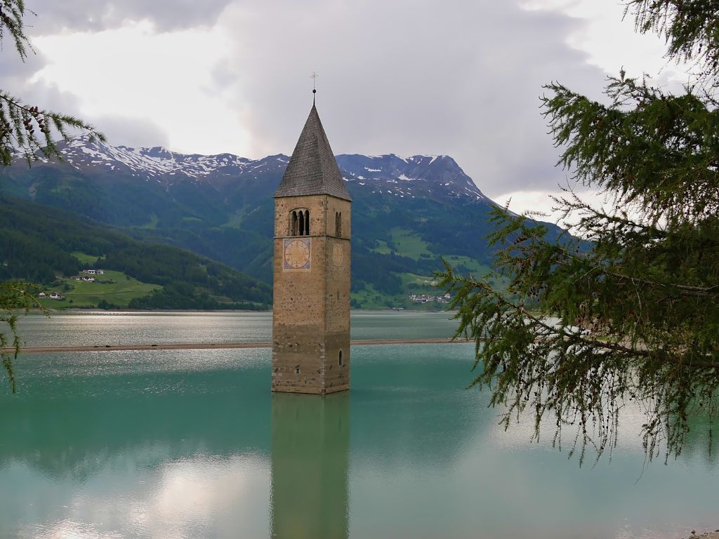 A stone church tower rises from the middle of a turquoise lake, with mountains and cloudy skies in the background, all framed by tree branches – one of the spookiest boating destinations in Europe.