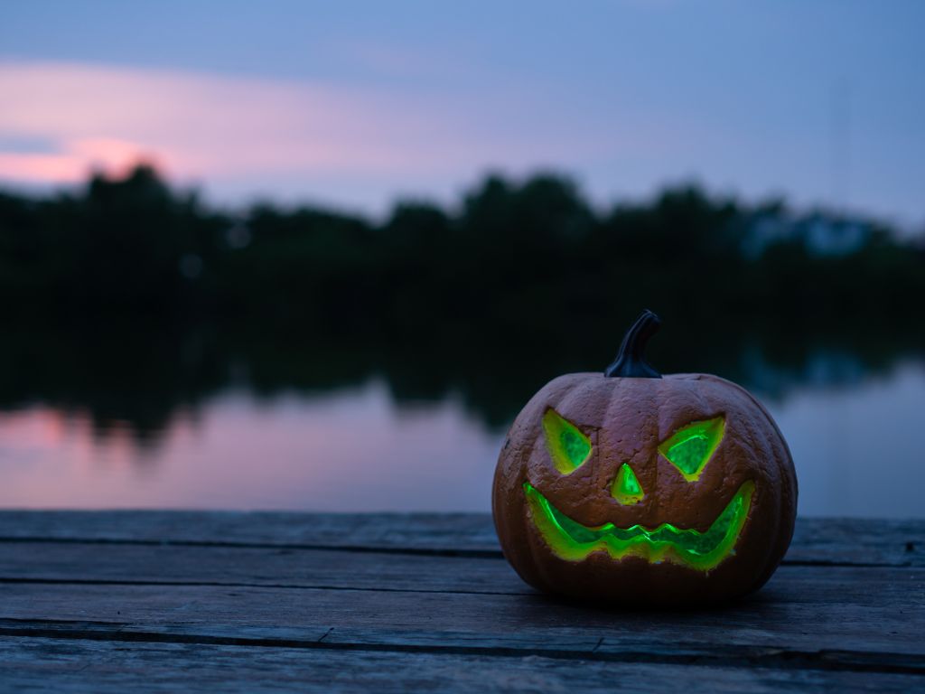 A carved pumpkin with a glowing green light inside sits on a wooden surface by a lake at dusk.