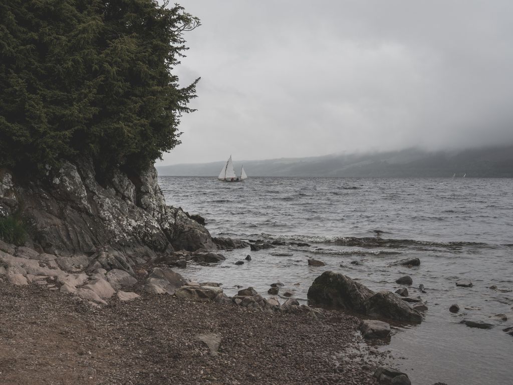 Rocky shoreline of Loch Ness with trees on the left, choppy water, and two sailboats in the distance under a cloudy, overcast sky.