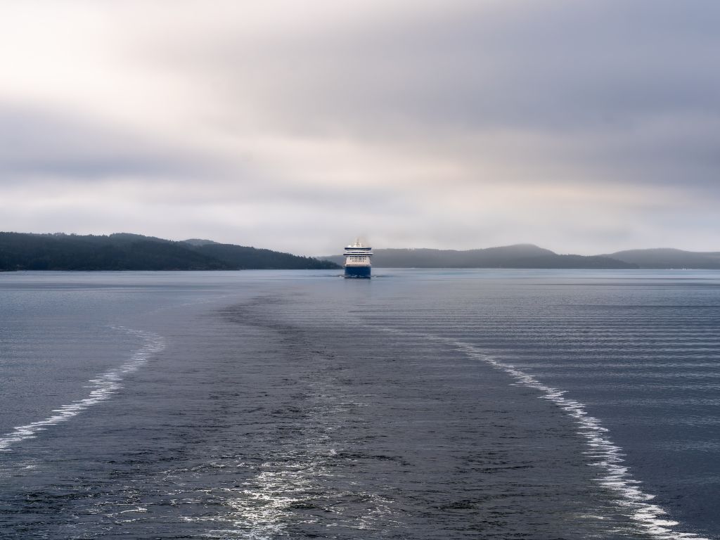 A distant view of a large ferry moving away across a calm body of water, leaving a visible wake beneath a cloudy sky with distant hills on the horizon.