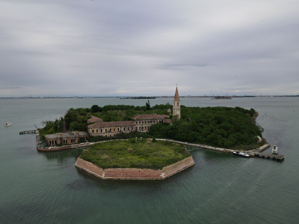 A small, spooky looking green island with historic buildings and a tall bell tower, all surrounded by water under a cloudy sky.