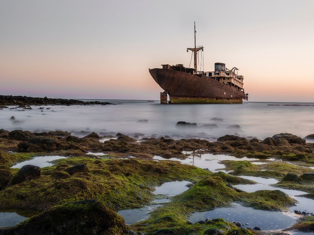 A rusted, abandoned shipwreck sits in shallow water near a rocky, moss-covered shoreline under a clear sky at sunset. Spooky looking ship.