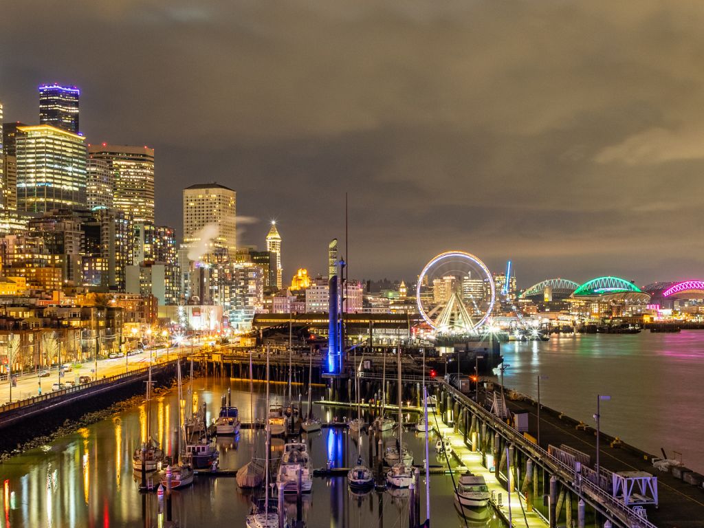 Seattle' waterfront at night, with glowing buildings, a Ferris wheel, festive marina boats, and bridges lit green and pink reflecting on the water.