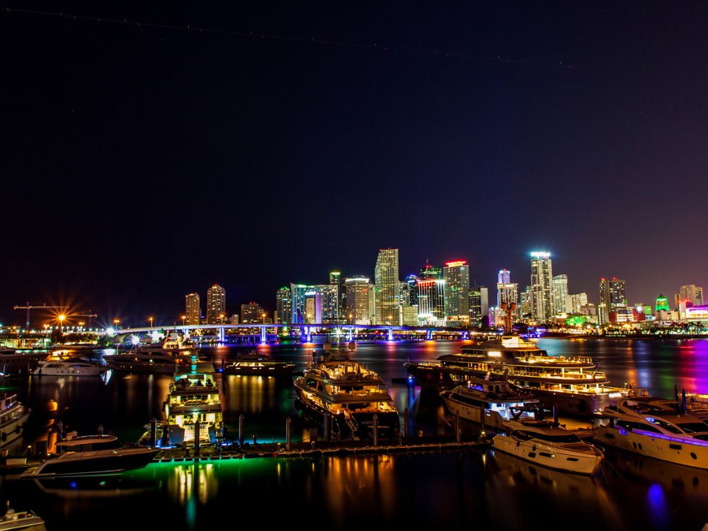 A marina with docked boats at night, ideal for a Christmas boating trip, with Miami's city skyline lit by colorful lights shimmering on the water.