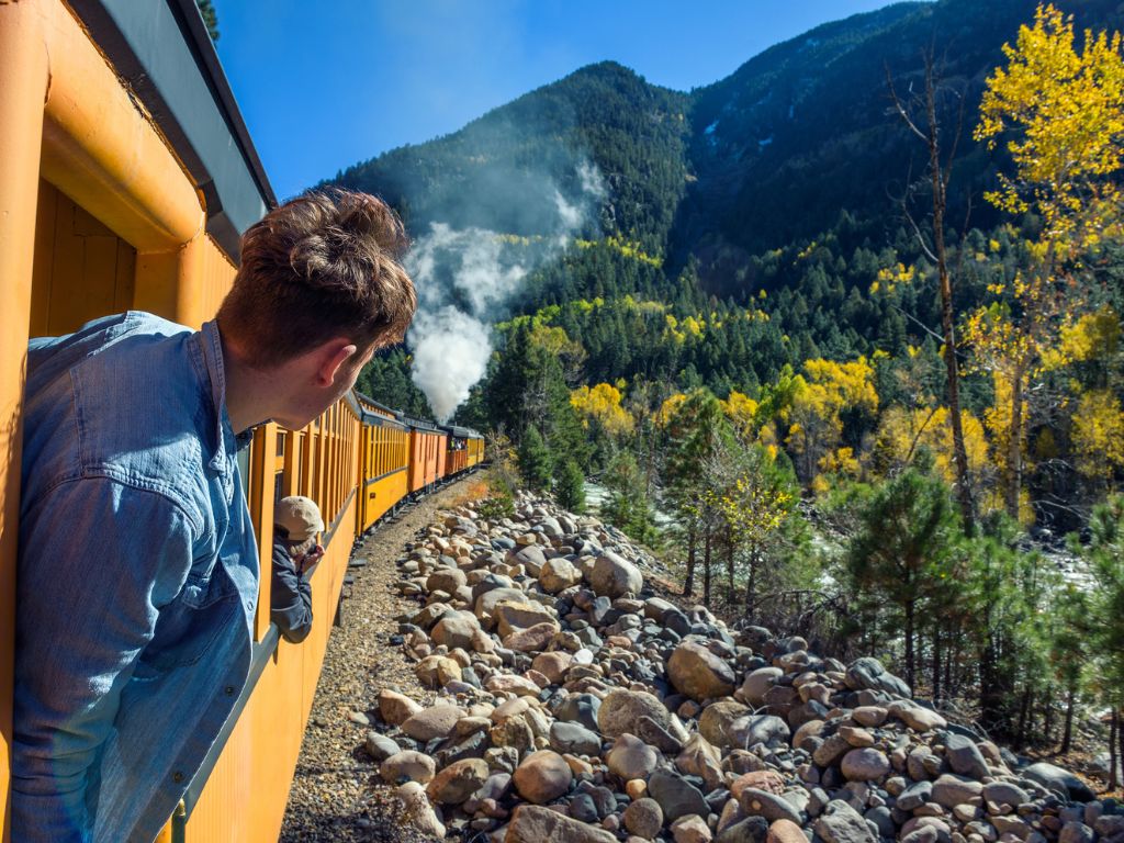 A man leans out of a yellow train car as it travels through a rocky mountain landscape, among trees and distant smoke.