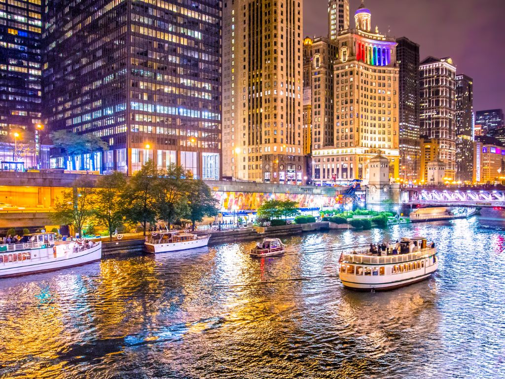 Boats cruise along a brightly lit river in Chicago at night, making this city's skyline a magical boating spot with towers and festive lights reflected on the water.