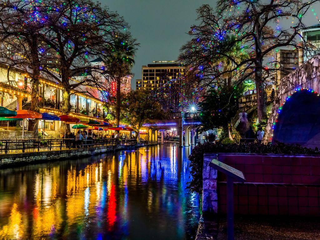It’s a nighttime view of a riverwalk, a charming Christmas boating spot lined with colorful lights, umbrella-covered restaurant tables, and shimmering reflections on the water.