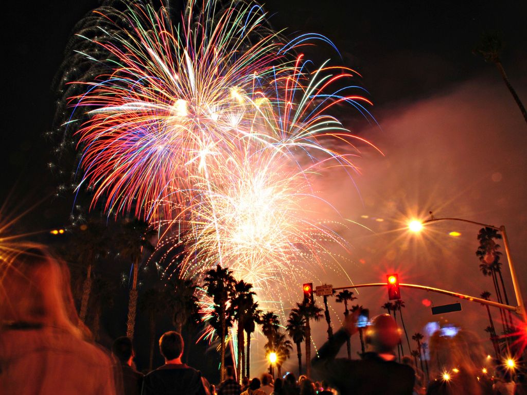 A crowd watching colorful fireworks explode in the night sky above palm trees, celebrating at a popular Christmas boating spot with illuminated traffic lights.