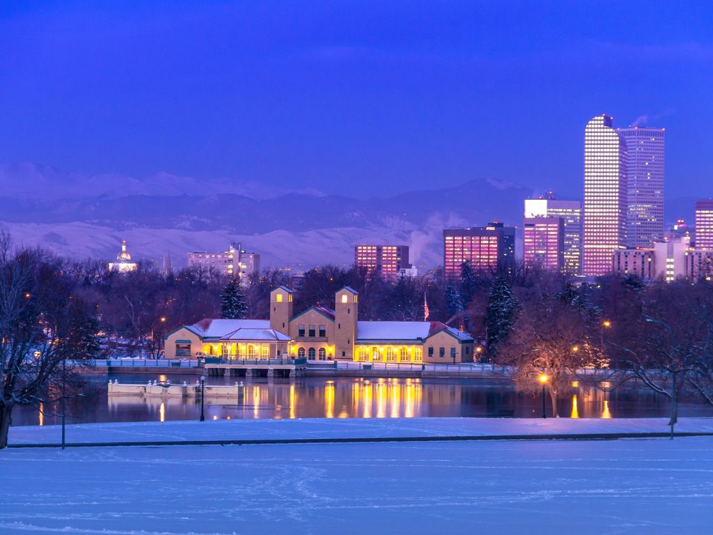 A snow-covered park with a lake up front, an illuminated building by the water, and a city skyline – all making a magical Christmas boating spot at twilight.