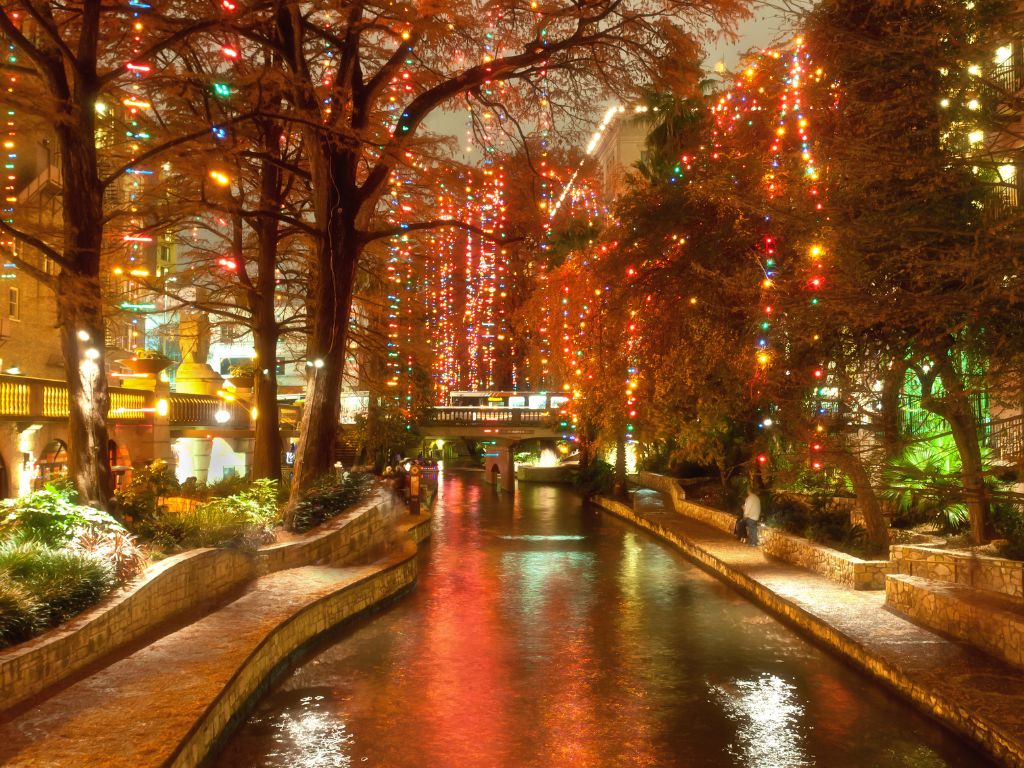 A canal lined with trees and buildings is lit up with colorful holiday lights at night, reflecting on the water as people stroll along this charming Christmas boating spot.