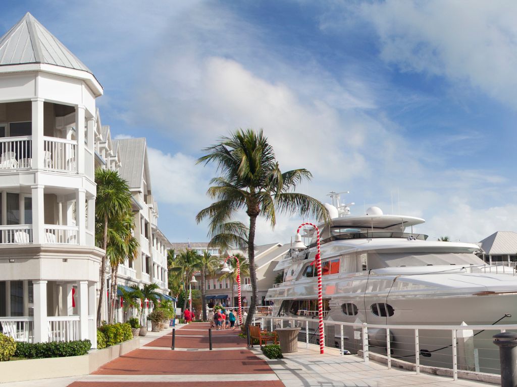 A marina walkway in Key West lined with palm trees, white buildings with balconies, and large yachts docked under a partly cloudy sky.