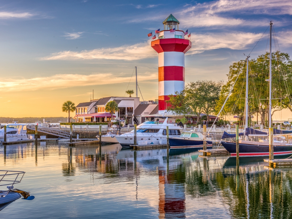 The Harbour Town Lighthouse, overlooking a calm Hilton Head marina at sunset, with sailboats and yachts docked along the waterfront.