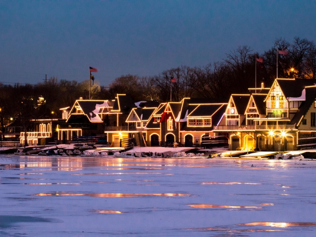 Philadelphia's waterfront at night in winter, with boathouse-style buildings outlined in holiday lights reflecting on the icy river.