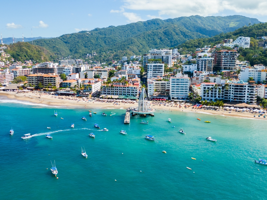 An aerial view of Puerto Vallarta’s waterfront with the turquoise Banderas Bay, boats scattered across the water, sandy beach, and the city backed by green mountains.