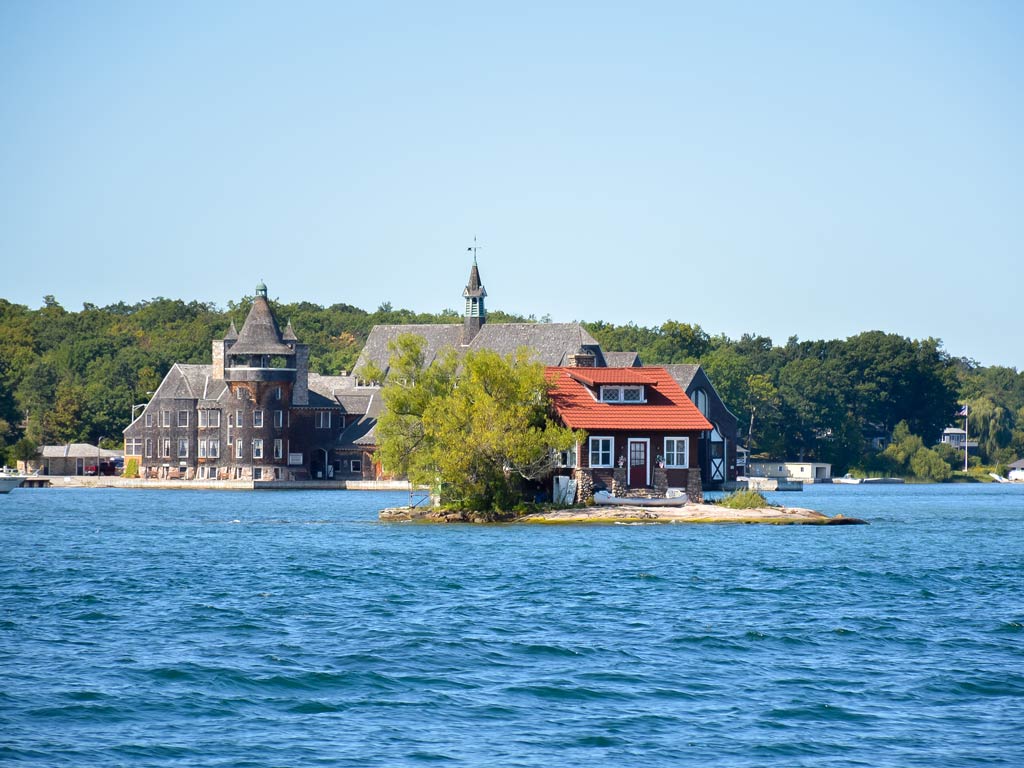 There's a small house with a red roof on a tiny island surrounded by blue water with larger buildings and trees rising in the background.