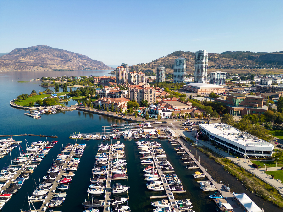 Here's an aerial view of a marina in Kelowna with boats docked, modern buildings, and mountains in the background, under a clear sky.