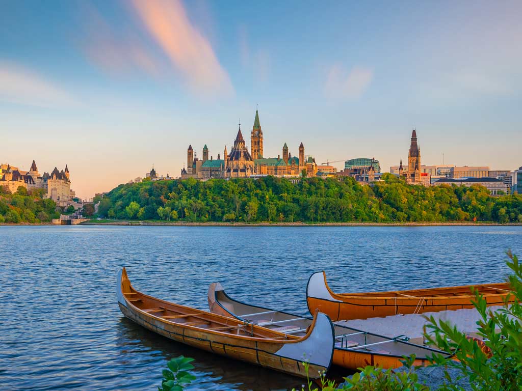 Two wooden canoes float on the Ottawa River with Ottawa’s Parliament Hill in the background, showing why this city’s one of the best boating towns in Canada.