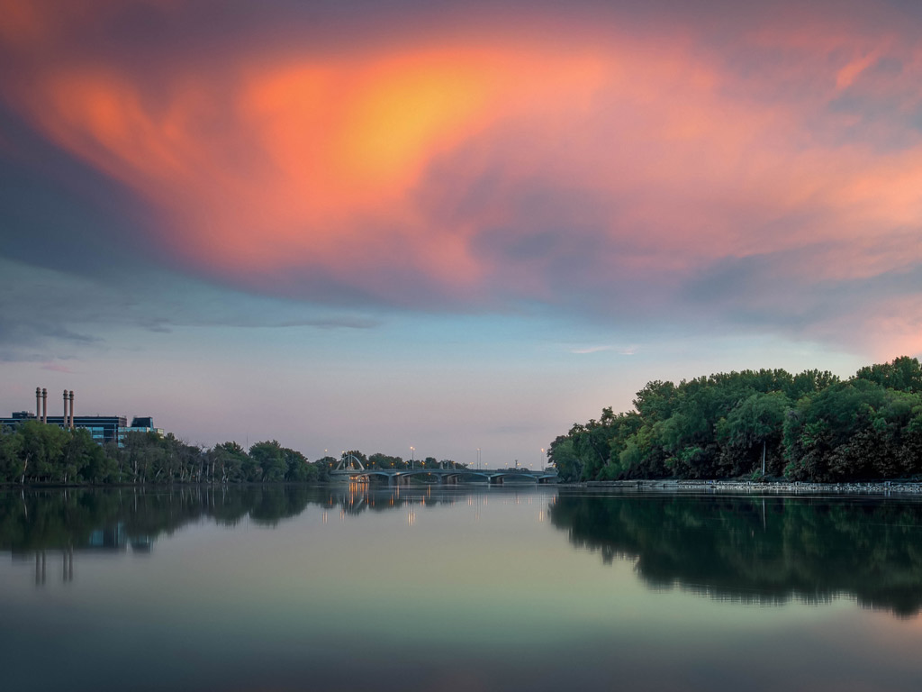 There’s a calm river with green trees on both banks, a distant bridge, and an orange evening cloud—classic scenery from one of the Best Boating Towns in Canada.