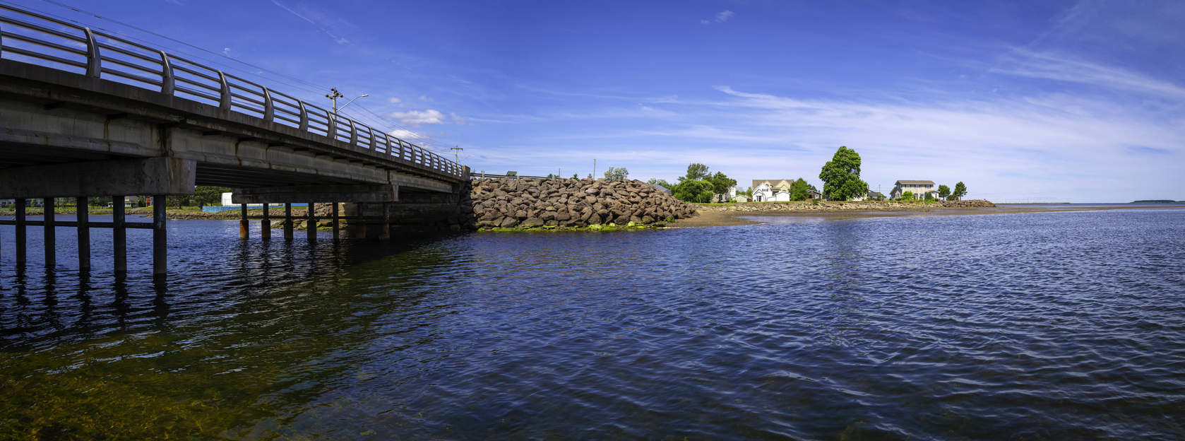 A bridge stretches over blue water toward a rocky shore with buildings and trees, showing the charm of one of Canada’s Best Boating Towns beneath a clear blue sky.