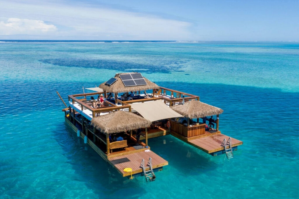 A floating bar with thatched roofs and solar panels offers unique boat dining on clear turquoise water, with Coral Reefs and open ocean all around under a blue sky.