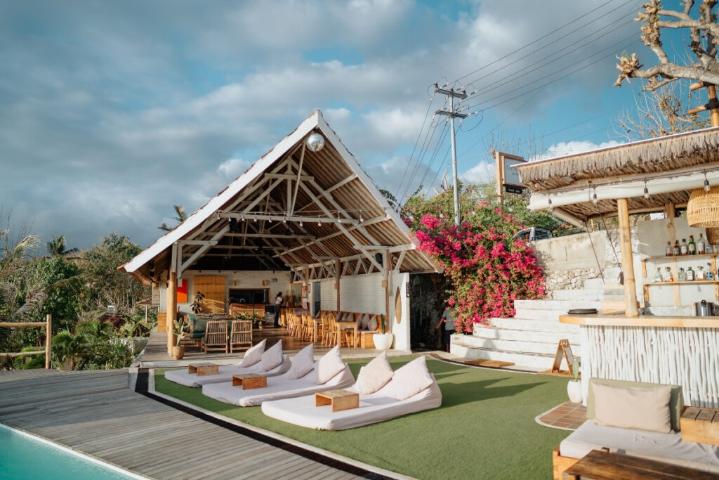 Chill Penida Restaurant - outdoor poolside lounge area with white cushioned floor seats, a thatched roof bar, and open-air wooden structure; enjoy boat dining amid greenery and flowers under a cloudy sky.