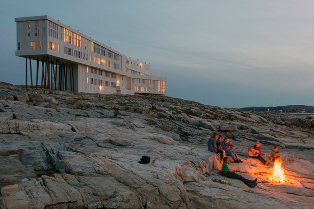 A group of people're sitting around a campfire on rocky terrain near a modern stilted building with many windows at dusk, swapping stories of boat dining adventures, including Cobia and Red Snapper feasts.