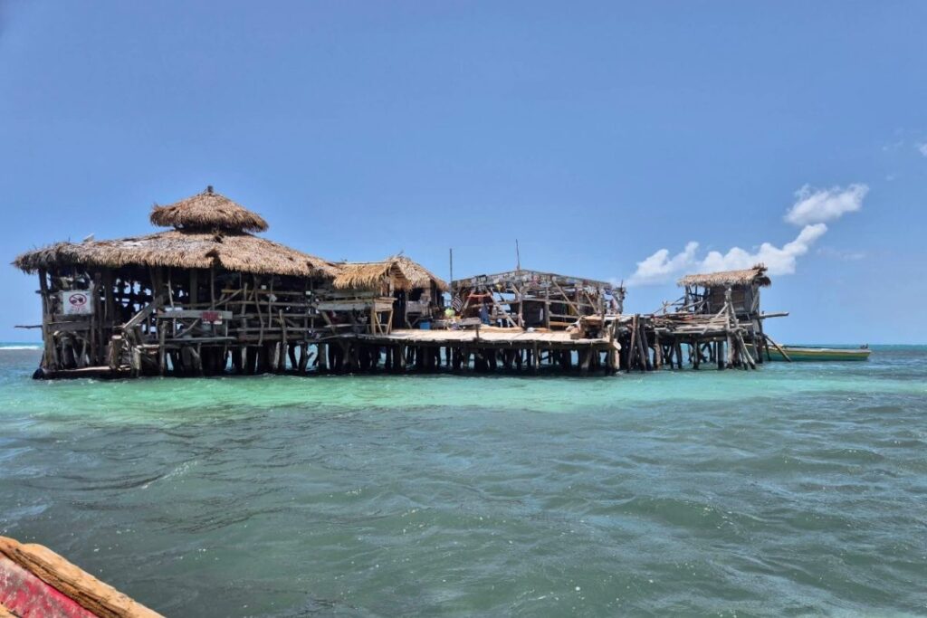 A rustic wooden floating bar with thatched roofs stands on stilts above clear blue ocean water, offering unique boat dining under a bright sky.