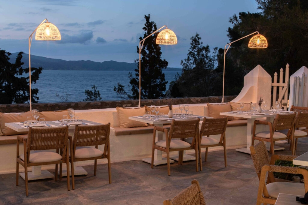 You’ll find an outdoor restaurant patio with wicker chairs, set tables, and rattan lamps that’s offering boat dining views of the sea at dusk, with trees and a white wall all around.