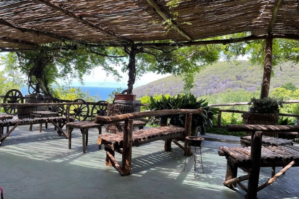Outdoor terrace with rustic wooden benches and tables under a wooden pergola, perfect for boat dining while overlooking green hills and the blue sea in the distance.