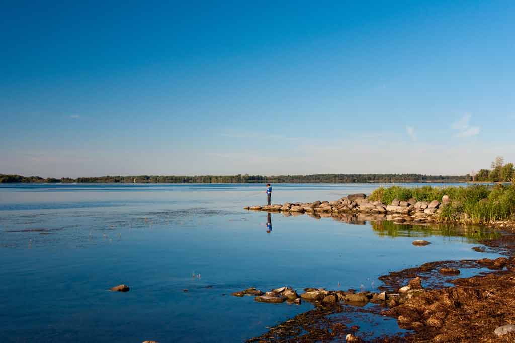 Someone's standing on a rocky shore by a calm lake under a clear blue sky, enjoying one of the Best Boating Towns in Canada. You can see distant trees along the horizon.