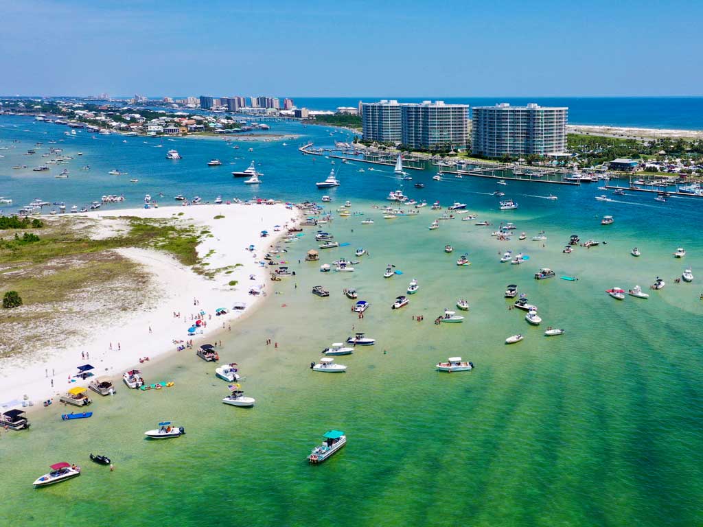 Aerial view of Memorial Day boating with plenty of boats in shallow turquoise water, people on the sand, and several high-rise buildings in the background.