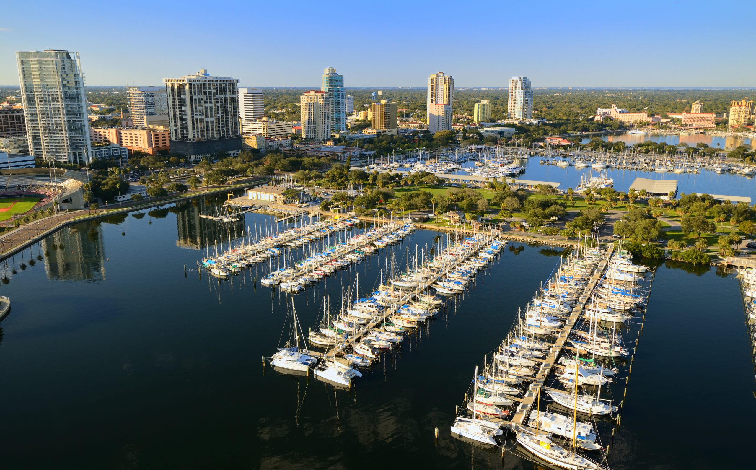 You’re looking at an aerial view of a marina with several docks of boats and yachts—set against high-rise city buildings and a clear blue sky.