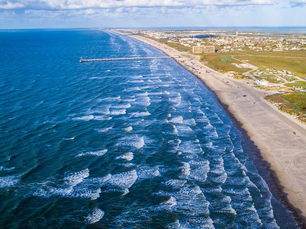 You’re looking down at a sandy beach with gentle waves and boats near a long pier, and buildings along the shore under a partly cloudy sky.