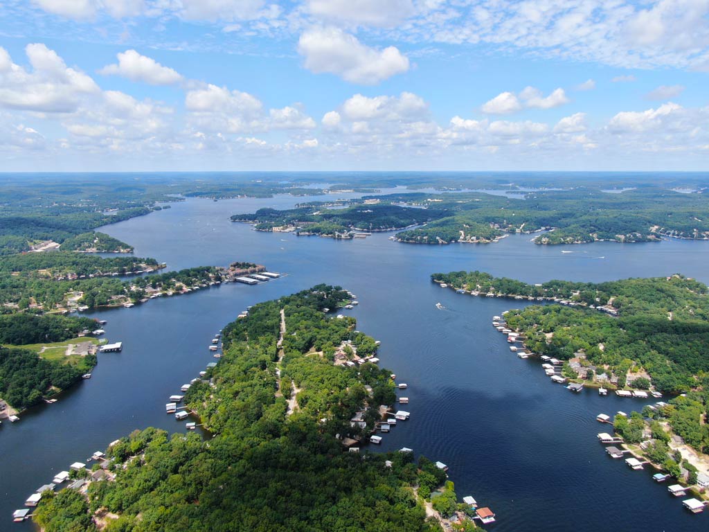 You’re looking at an aerial view of a big lake with tree-covered peninsulas, docks, and scattered homes beneath a partly cloudy sky. 